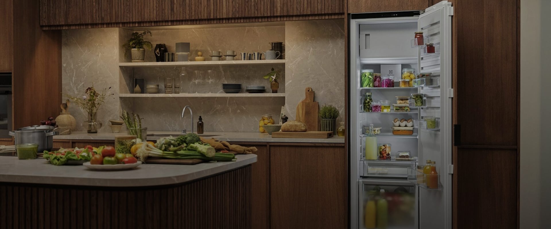 Overblik over et åbent, integreret Electrolux køleskab i et moderne køkken, fyldt med friske råvarer, flasker og organiserede bøtter.